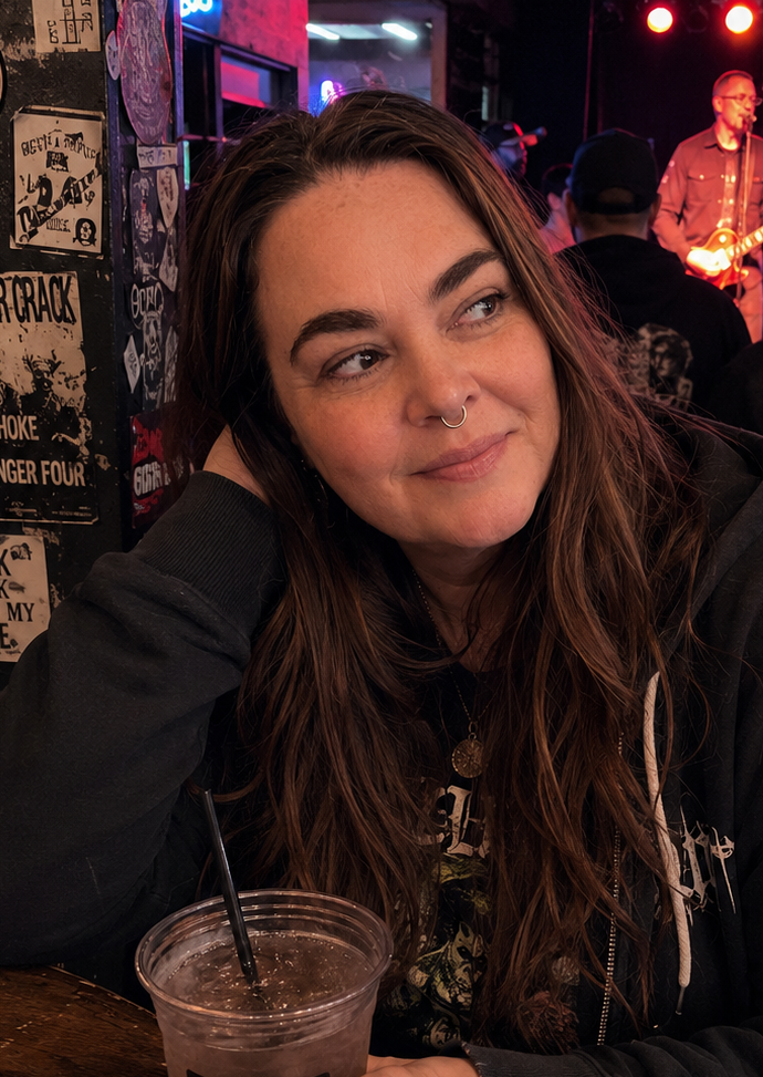 Nicole at Reverb in Reading, PA, leaning on a sticker-covered pillar with an iced coffee while Leftover Crack plays on stage behind her.